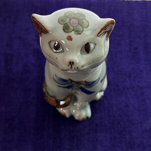 May include: A ceramic cat figurine with a pale blue glaze. The cat has brown-tipped ears, dark eyes, and a floral design on its head. Blue ribbon details adorn the chest. A decorative item.