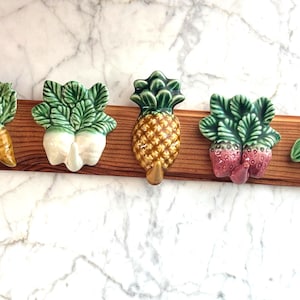 May include: A wooden plaque with five ceramic vegetable-shaped hooks. The hooks depict carrots, radishes, a pineapple, strawberries, and corn. The vegetables are painted in bright colours, with green leaves and a brown wooden backing.