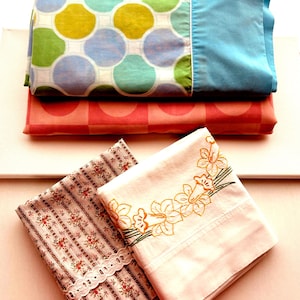 May include: A selection of folded pillowcases in different patterns and colours. The top pillowcase is blue, with a white border. Below it is a pillowcase with large, colourful circles. The bottom pillowcases have floral and embroidered designs.