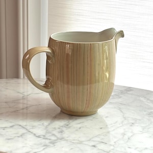May include: A ceramic jug featuring a glossy finish with vertical stripes in shades of beige, soft green, and muted orange. The jug has a curved handle on the side, a rounded body, and a distinct pouring spout at the top rim.