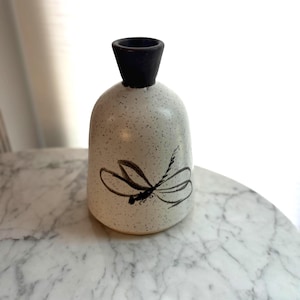 May include: A small ceramic bud vase with a speckled off-white body and a dark brown, flared neck. The front of the vase features a hand-painted black dragonfly illustration with delicate wing details and a slender body, centred on the rounded base.