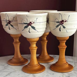 May include: A set of four goblets featuring off-white ceramic bowls with horizontal ridges and a hand-painted holly berry and leaf motif. Each bowl is supported by a turned light brown wooden stem and a circular wooden base.