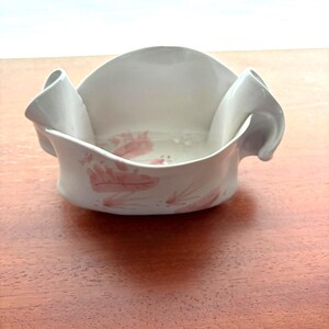 May include: A white ceramic bowl with a ruffled rim and two curved handles. The bowl has a delicate pink floral pattern on the interior and exterior. The bowl is on a wooden surface.