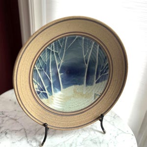 May include: A round ceramic plate with a speckled tan rim featuring a dark blue and white painted forest scene in the centre. The design depicts stylised white tree trunks against a deep blue background, with a light tan foreground area.