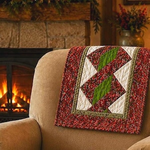 May include: A quilted throw blanket featuring a central panel of white and bright green diamond shapes surrounded by a red fabric border with a detailed floral pattern. The quilt has visible stitching throughout and is draped over a tan armchair.