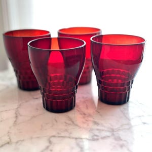 May include: Four ruby red drinking glasses with a geometric design. The glasses have a flared shape and are made of a translucent material. The glasses are arranged on a marble surface.