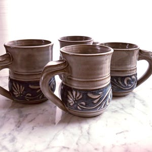 May include: A set of four ceramic mugs featuring a two-tone design with a smooth, light beige upper section and a dark blue lower section decorated with embossed floral patterns. Each mug has a wide, curved handle and a sturdy, rounded base.