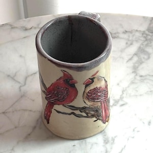 May include: A ceramic mug with a cream-coloured exterior featuring a hand-painted illustration of two cardinal birds perched on a brown branch. The mug has a dark grey interior and a dark grey rim, with a sturdy handle extending from the side.
