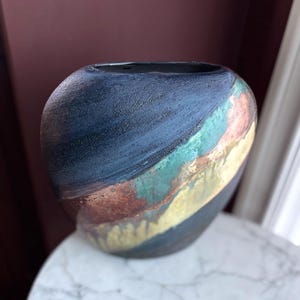 Peut inclure: Un vase en céramique fait à la main avec un extérieur bleu foncé et une bande diagonale de couleurs comprenant du vert, de l'or et du cuivre. Le vase a une surface texturée et une forme arrondie, adapté à la décoration intérieure.