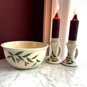 May include: A cream-coloured ceramic bowl and two matching candle holders, each featuring a hand-painted design of green leaves and small red berries. Two lit dark red pillar candles are placed inside the ceramic candle holders.