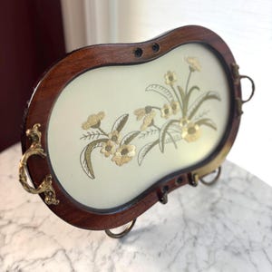 May include: An oval wall-mounted key holder with a dark wood frame and a cream-coloured centre panel featuring embroidered gold floral designs. The piece includes ornate brass side handles and three small brass hooks along the bottom edge.
