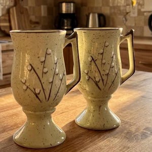 Peut inclure: Deux grandes tasses en céramique sur pied avec un émail jaune pâle moucheté. Chaque tasse présente un motif botanique peint à la main en brun foncé, composé de tiges avec des bourgeons blancs en relief. Elles ont un bord évasé et une anse.
