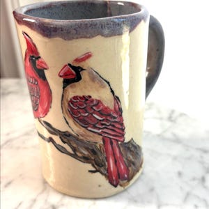 May include: A ceramic mug with a cream-coloured glaze featuring a hand-painted illustration of two northern cardinal birds perched on a brown tree branch. The mug has a dark, mottled purple-grey rim and a matching dark handle on the right side.