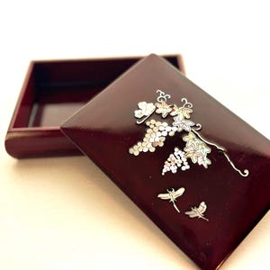May include: A rectangular, claret-coloured wooden box with a hinged lid. The lid is decorated with an inlaid design of grapes, leaves, and dragonflies. The box is open, revealing the interior.