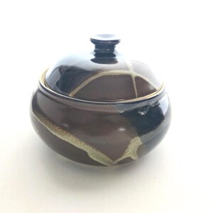 May include: A ceramic lidded jar in dark brown with a pattern of light tan and black streaks. The lid has a small, dark knob. The jar is likely for storage or decoration. The jar's measurements are in centimetres.