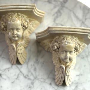 May include: A pair of decorative wall shelves, each with a cherubic face and detailed wings. The shelves are a creamy beige colour and have an ornate design. They are likely made of a durable material and are suitable for displaying small items.
