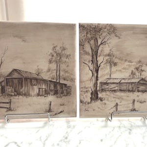 May include: Two square ceramic tiles featuring monochromatic sepia-toned illustrations of rural barn scenes. Each tile depicts a wooden barn structure, a fence, and trees set against a cloudy sky. The artwork is rendered in a sketch-like style.