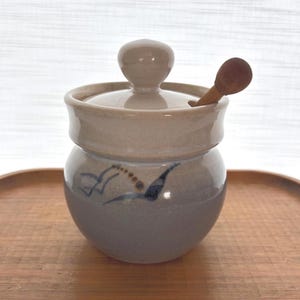 May include: A ceramic sugar bowl with a lid and a small wooden spoon. The bowl is two-toned, with a light beige top and a grey-blue base. A blue bird design decorates the side. The bowl sits on a wooden tray.