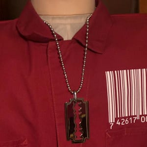 May include: A silver-colored pendant necklace featuring a razor blade design. The necklace hangs on a ball chain. The background is a red shirt with a barcode graphic and the numbers 7 42617 00.