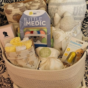 May include: A baby gift basket filled with various items. Includes a plush bear, a book titled "Little Medic", a finger toothbrush, washcloths, and other baby essentials. The basket is made of woven material.