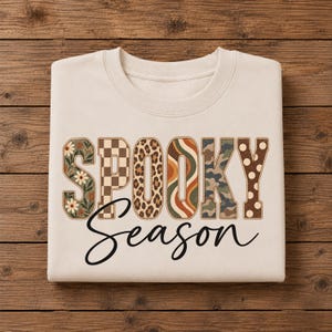Könnte beinhalten: Ein beigefarbenes Sweatshirt mit Rundhalsausschnitt und dem Schriftzug SPOOKY SEASON. Das Wort SPOOKY ist in großen Blockbuchstaben gedruckt, wobei jeder Buchstabe ein einzigartiges Muster wie Blumen, Karos, Leopardenmuster, Wellenstreifen und Punkte aufweist.