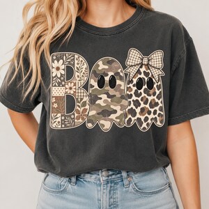 以下が含まれることがあります： 大きな文字で「BOO」とプリントされたチャコールグレーの半袖Tシャツ。Bは花柄とチェック柄、1つ目のOは迷彩柄のゴースト、2つ目のOはギンガムチェックのリボンをつけたヒョウ柄のゴーストのデザインです。