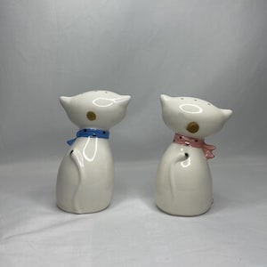 May include: Two white ceramic cat-shaped salt and pepper shakers. Each shaker has a small, round gold accent and a colorful scarf. One cat has a blue scarf, and the other has a pink scarf.