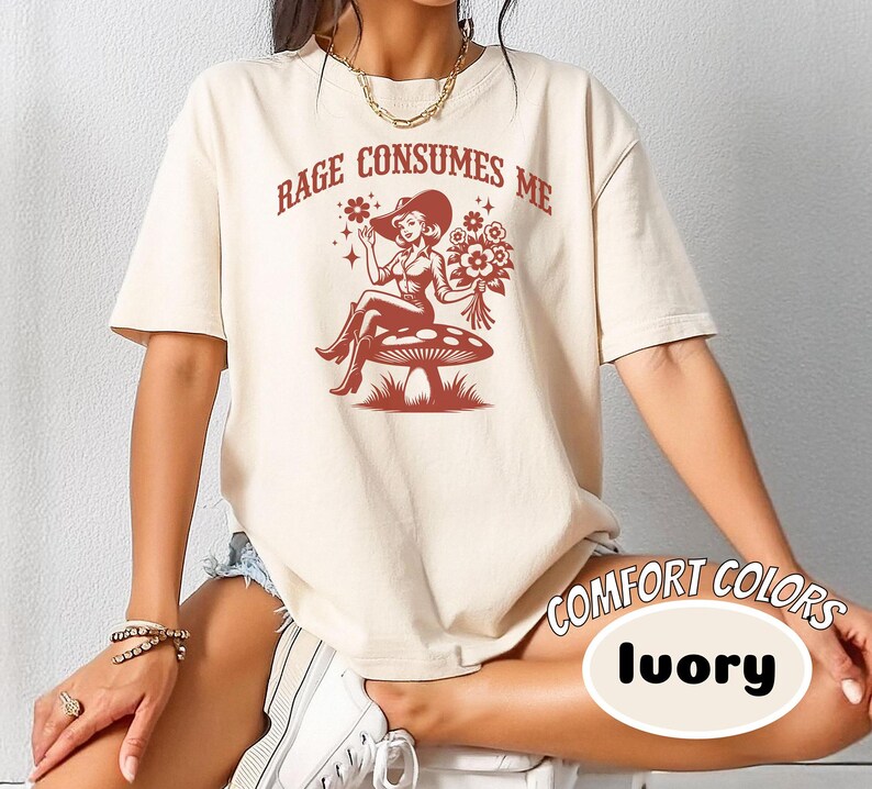 K&ouml;nnte beinhalten: Ein cremefarbenes T-Shirt mit einem Aufdruck einer Frau in einem Cowboyhut, die auf einem Pilz sitzt. Der Text "Rage Consumes Me" ist auf dem Shirt gedruckt.