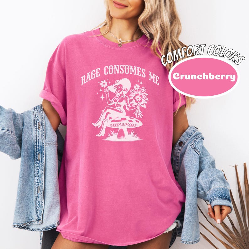 K&ouml;nnte beinhalten: Ein pinkfarbenes T-Shirt mit dem Text "Rage Consumes Me" und einer wei&szlig;en Illustration einer Frau mit Hut, die auf einem Pilz sitzt.