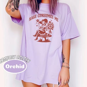 K&ouml;nnte beinhalten: Ein lavendelfarbenes T-Shirt mit dem Aufdruck "Rage Consumes Me" in Braun. Die Grafik zeigt eine Frau im Cowboyhut, die auf einem Pilz mit Blumen sitzt.
