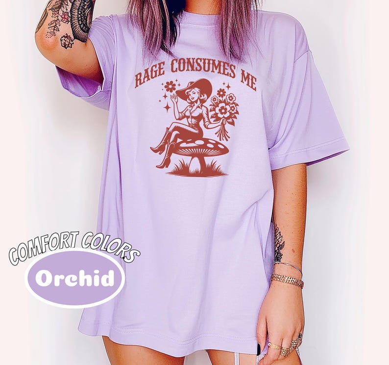 K&ouml;nnte beinhalten: Ein lavendelfarbenes T-Shirt mit dem Aufdruck "Rage Consumes Me" in Braun. Die Grafik zeigt eine Frau im Cowboyhut, die auf einem Pilz mit Blumen sitzt.