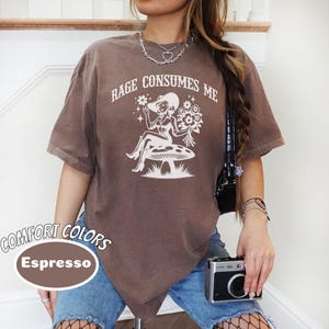 K&ouml;nnte beinhalten: Ein braunes T-Shirt mit dem Text "Rage Consumes Me" und einer Grafik einer Frau mit Hut, die auf einem Pilz sitzt. Das Shirt ist aus einem weichen, bequemen Material und hat einen Used-Look.