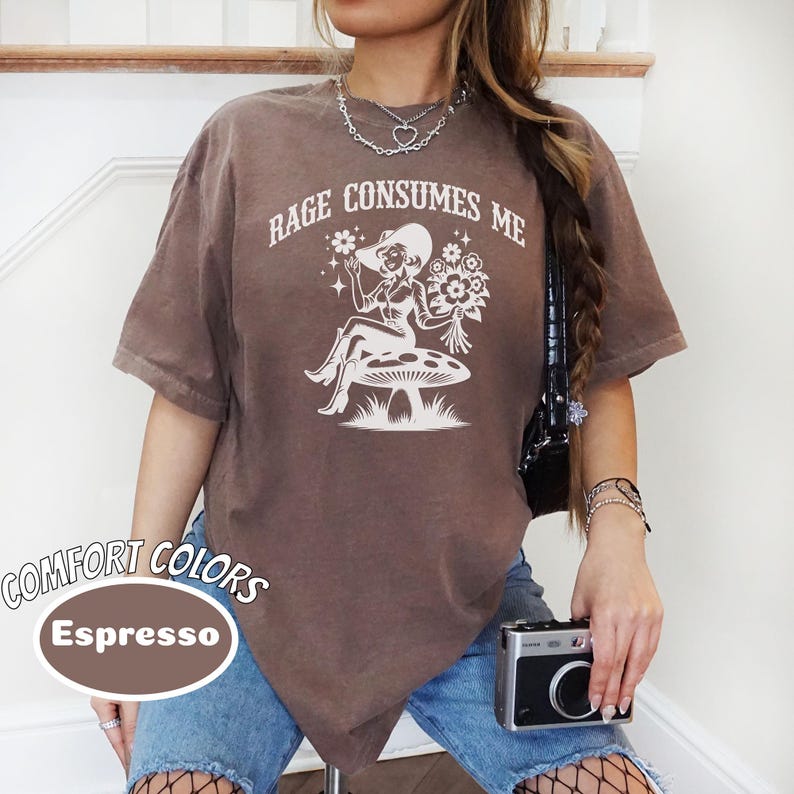 K&ouml;nnte beinhalten: Ein braunes T-Shirt mit dem Text "Rage Consumes Me" und einer Grafik einer Frau mit Hut, die auf einem Pilz sitzt. Das Shirt ist aus einem weichen, bequemen Material und hat einen Used-Look.