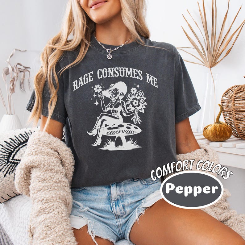 K&ouml;nnte beinhalten: Ein dunkelgraues T-Shirt mit einem wei&szlig;en Grafikdruck einer Frau, die auf einem Pilz mit Blumen sitzt. Der Text "Rage Consumes Me" ist in Wei&szlig; auf das Shirt gedruckt. Das Shirt ist Teil der Marke Comfort Colors und hat die Farbe Pepper.