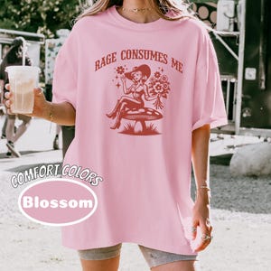 K&ouml;nnte beinhalten: Rosa &uuml;berdimensioniertes T-Shirt mit einer roten Grafik einer Frau, die auf einem Pilz sitzt, mit dem Text "Rage Consumes Me".