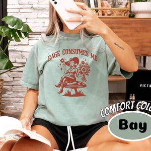 K&ouml;nnte beinhalten: Ein blaugr&uuml;nes T-Shirt mit einem Aufdruck einer Frau in einem roten Kleid und einer Blumenkrone, die auf einem Pilz sitzt. Der Text "Rage Consumes Me" ist in Rot auf dem Shirt gedruckt.