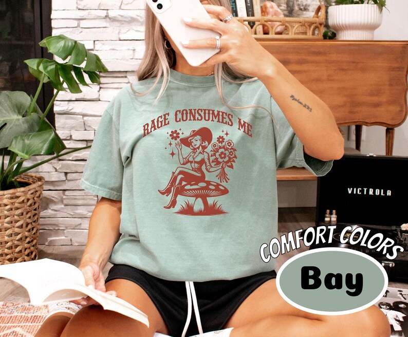 K&ouml;nnte beinhalten: Ein blaugr&uuml;nes T-Shirt mit einem Aufdruck einer Frau in einem roten Kleid und einer Blumenkrone, die auf einem Pilz sitzt. Der Text "Rage Consumes Me" ist in Rot auf dem Shirt gedruckt.
