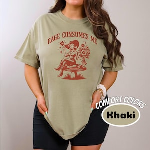 K&ouml;nnte beinhalten: Ein khakifarbenes T-Shirt mit dem Text "Rage Consumes Me" und einer Grafik einer Frau in einem Cowboyhut, die auf einem Pilz sitzt. Die Grafik ist in Rot und der Text ist in Rot.