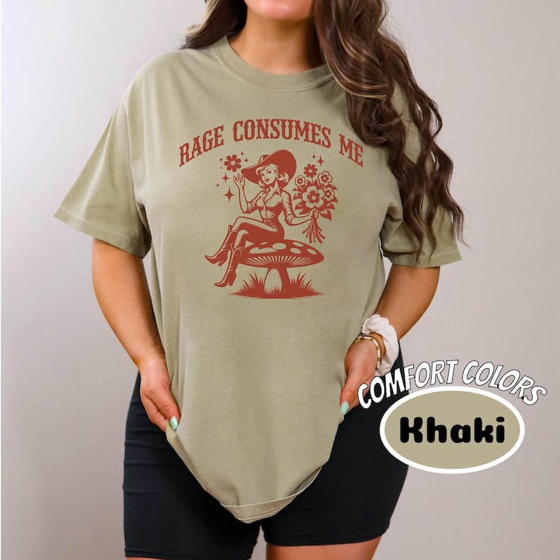 K&ouml;nnte beinhalten: Ein khakifarbenes T-Shirt mit dem Text "Rage Consumes Me" und einer Grafik einer Frau in einem Cowboyhut, die auf einem Pilz sitzt. Die Grafik ist in Rot und der Text ist in Rot.