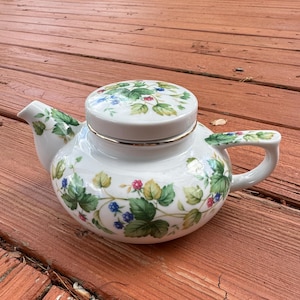 May include: A white ceramic teapot featuring a botanical pattern of green leaves, blue berries, and small red berries. The teapot has a matching lid with a gold-tone rim, a curved handle, and a short spout, all decorated with the same berry motif.