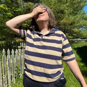 May include: A short-sleeved polo shirt featuring horizontal stripes in navy blue and tan. The garment has a classic collar, a buttoned placket at the neckline, and a relaxed fit. The fabric displays a consistent striped pattern across the chest and sleeves.