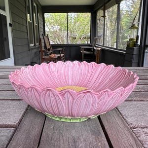 Op de afbeelding: Een grote, roze keramische kom in de vorm van een bloem. De kom heeft een gele kern en een groene basis. De kom staat op een houten tafel op een veranda.