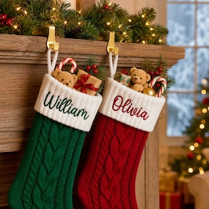 Op de afbeelding: Twee gebreide kerstsokken hangend aan gouden haken aan een houten schoorsteenmantel. De linker sok is groen met de naam William in het groen geborduurd, en de rechter is rood met de naam Olivia in het rood geborduurd. Beide hebben witte boorden.