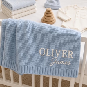 May include: A light blue knit baby blanket featuring the embroidered name OLIVER James in white thread. The blanket has a ribbed border and is displayed alongside a stack of folded blankets, a light blue beanie, a wooden stacking toy, and a white bodysuit.
