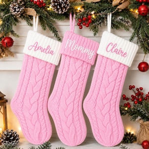 Puede incluir: Tres calcetines navideños de punto rosa con puños acanalados blancos. Cada calcetín presenta un patrón de trenzas y está personalizado con nombres bordados en letra cursiva rosa: Amelia, Mommy y Claire, colgados de un lazo blanco.