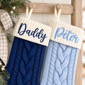 May include: Two cable-knit Christmas stockings with cream-colored ribbed cuffs. The left stocking is dark navy blue with the name Daddy embroidered in dark blue. The right stocking is light blue with the name Peter embroidered in light blue.