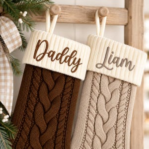 Peut inclure: Deux chaussettes de Noël en tricot torsadé suspendues à un support en bois. La chaussette de gauche est marron foncé avec le mot Daddy brodé en marron. Celle de droite est beige avec le mot Liam brodé en marron. Toutes deux ont des revers crème.