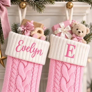 Op de afbeelding: Twee roze kerstsokken van kabelgebreid materiaal met witte boorden. De linker sok is geborduurd met de naam Evelyn in roze schrijfletters. De rechter sok is geborduurd met de letter E in het roze. Beide sokken zijn gevuld met cadeaus en speelgoed.