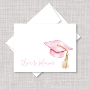 May include: A white folded note card and matching envelope featuring a watercolor illustration of a light pink graduation cap with a gold tassel. The name Olivia Williams is printed in a light pink script font on the lower left side of the card.