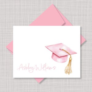 May include: A white rectangular greeting card featuring a watercolor illustration of a light pink graduation cap with a gold tassel. The name Ashley Williams is printed in a light pink script font on the lower left side of the card.